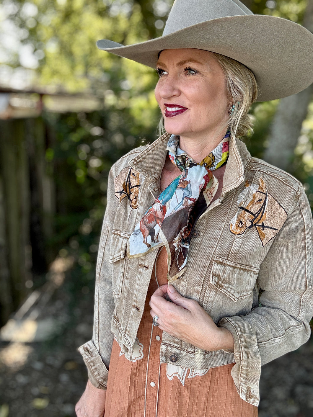 Woman wearing a beige cowboy hat and jacket with horse designs outdoors.