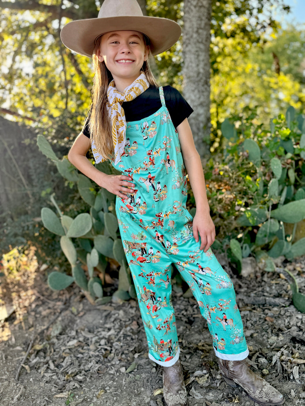 Child wearing a turquoise cowboy-themed jumpsuit with a hat and scarf, standing in front of cacti.