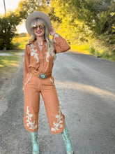 Woman in a peach shirt and pants set with white embroidery standing on a road with greenery in the background