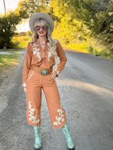 Woman in a vintage-style outfit with a cowboy hat, sunglasses, and turquoise boots standing on a road.