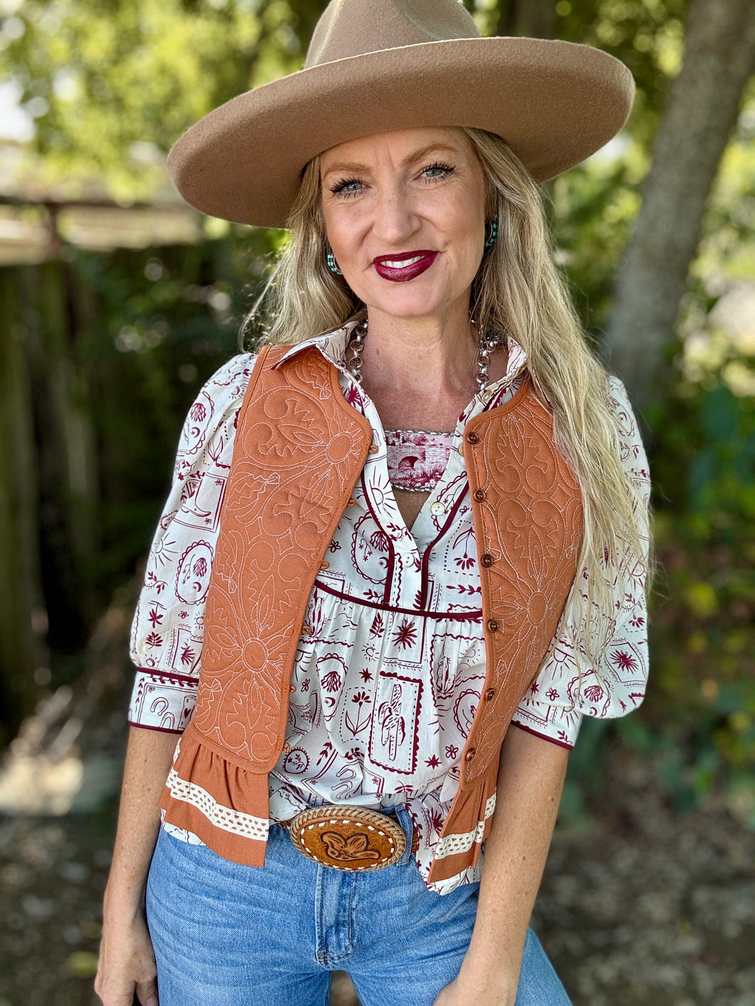 Woman wearing a cowboy hat, patterned shirt, vest, and jeans outdoors.