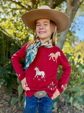 Child wearing a red sweater with horse designs, a cowboy hat, and a scarf outdoors.