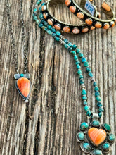 A styled flat-lay of bohemian jewelry on a weathered wood background, featuring a silver-tone necklace with a coral-orange stone pendant, turquoise bead strands, matching turquoise and coral bracelets, and a matching turquoise and coral pendant.