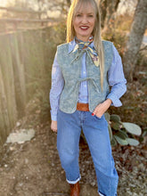 Woman wearing a blue shirt, patterned vest, and jeans outdoors with trees and rocks in the background.