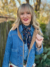 Woman wearing a blue denim jacket with a floral patterned scarf and dress, standing outdoors.