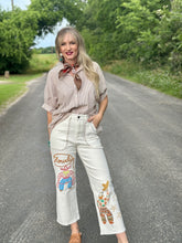 Woman standing on a road wearing a beige shirt and white pants with colorful designs.