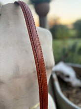 A close-up of a brown leather strap with embossed floral detailing and a braided edge, draped over a light beige fabric-covered display form against a softly blurred outdoor background.