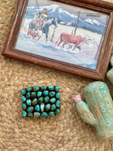A rectangular turquoise stone belt buckle with a silver-toned border, displayed on a woven jute mat next to a small cactus figurine and a framed western landscape print.