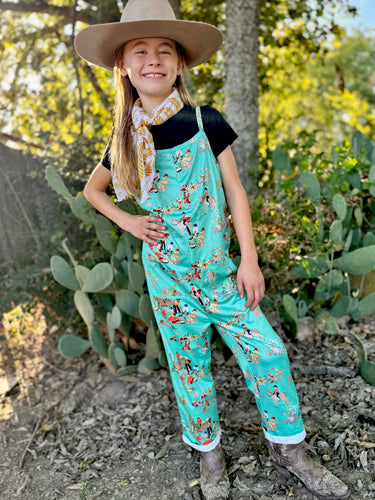 Child wearing a turquoise cowboy-themed jumpsuit with a hat and scarf, standing in front of cacti.