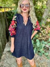 A woman wearing a dark indigo denim tunic dress with vibrant red, pink, and green floral embroidery on the sleeves, styled with brown boots and sunglasses in an outdoor garden setting.