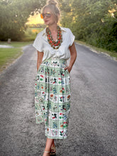 Woman in a white blouse and patterned skirt standing on a road with greenery in the background