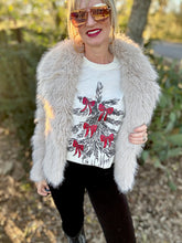 Woman wearing a white graphic t-shirt with red bows, black pants, and a light gray fur coat outdoors.