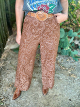 A person outdoors wearing brown paisley wide-leg pants with a matching brown belt and a large decorative buckle, paired with a gray graphic top and brown pointed shoes.