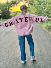 Person wearing a pink sweatshirt with 'GRATEFUL' on the back, standing on a road with trees and sunset in the background.