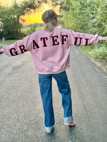 Person wearing a pink sweatshirt with 'GRATEFUL' on the back, standing on a road with trees and sunset in the background.