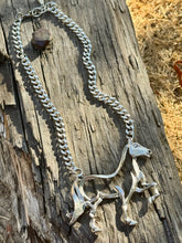 A silver-toned curb chain necklace with a sculptural horse pendant, displayed on a weathered wooden log outdoors.