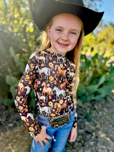 Young girl wearing a cowboy hat and patterned shirt with horses outdoors