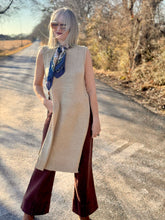 Woman standing on a road wearing a beige sleeveless sweater and brown pants with a blue scarf.