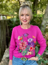 A woman wearing a bright pink crewneck sweater with a colorful floral and butterfly knit pattern, paired with blue jeans, standing outdoors in front of a blurred garden background.