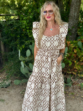 A woman wearing a bohemian-style tiered maxi dress with a brown geometric pattern on a cream background, standing outdoors in front of a garden with cacti.