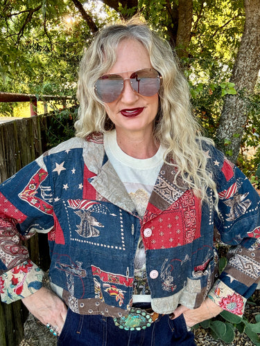 A woman with long blonde hair and sunglasses wearing a colorful patchwork denim jacket with patriotic American and western motifs, standing outdoors in front of trees and a wooden fence.
