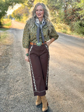 A woman stands on a gravel road outdoors wearing a bohemian-style outfit with a green patterned blouse, brown embroidered wide-leg pants, a blue and white patterned scarf, and tan boots.