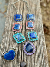 A handmade silver-toned jewelry set featuring multiple pieces with blue lapis lazuli stones in heart, teardrop, and square shapes, accented with pink, turquoise, and purple stones, displayed on a weathered wooden plank.