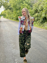 Woman wearing a colorful patchwork vest and camouflage pants on a road with greenery.
