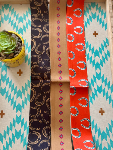 Three patterned twilly scarves on a wooden surface with a small plant in the corner.