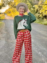 Person wearing a green sweater with a horse design and red plaid pants on a road with autumn foliage.