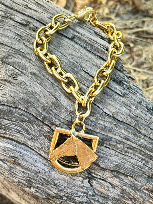 A gold chain bracelet with a shield-shaped horse head charm, displayed on a weathered gray wooden log outdoors.