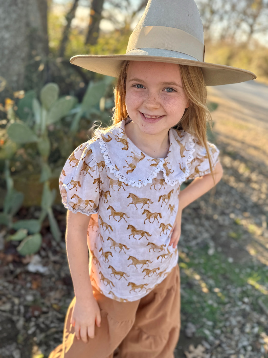 Young girl wearing a white shirt with animal prints and a wide-brimmed hat outdoors.