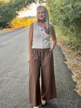 A woman stands on a paved country road wearing a sleeveless cream knit vest over a white top, brown wide-leg pinstriped trousers, and a red patterned scarf.