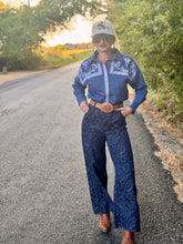 A woman stands on an outdoor paved road wearing a blue western-style long-sleeve shirt with white embroidery, matching dark blue patterned wide-leg pants, a large concho belt, and brown cowboy boots.