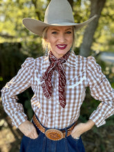 Woman wearing a cowboy hat, checkered shirt, and scarf with a blurred natural background