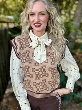 A woman with long wavy blonde hair wearing a beige knit sweater vest with a brown bow pattern, a cream polka-dot blouse with a large bow tie, and brown pants, standing outdoors in front of a blurred garden background.