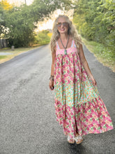 A woman wearing a pink tiered maxi dress with a multicolor floral print walks down a paved country road lined with green trees.