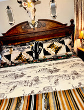 A styled bed in a bedroom featuring a western-themed bedding set with a cream quilted coverlet printed with black desert landscapes, a black cactus pillow, and a black, white, and gold geometric patterned shams, set against a dark wood headboard.
