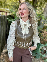 A woman wearing an olive faux-leather vest with white floral embroidery and pearl trim over a cream bow-tie blouse with small brown polka dots, paired with brown pants, photographed outdoors in front of a wooden fence and trees.