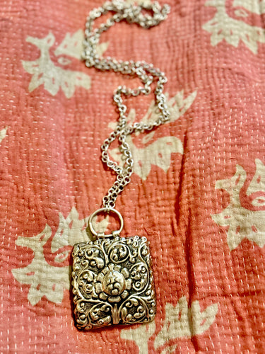 Decorative silver pendant on a chain against a floral-patterned fabric background