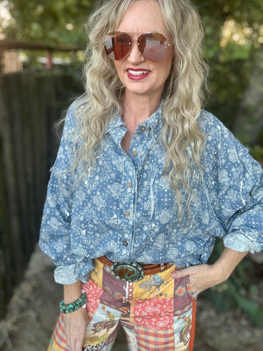 A woman wearing a light blue floral button-up shirt and colorful patchwork patchwork pants, styled with a large silver buckle belt and sunglasses, standing outdoors.