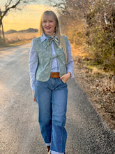 Woman standing on a rural road wearing a denim vest, blue jeans, and a brown belt.