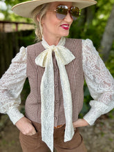 Woman wearing a checkered vest with a lace bow, white blouse, and brown pants outdoors.