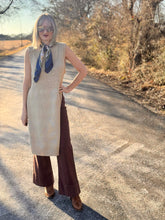 Woman standing on a road wearing a beige sleeveless dress and brown wide-leg pants.