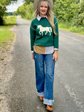 Woman wearing a green sweater with a horse design, blue jeans, and brown boots on a road.