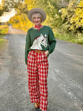 Woman wearing a sweater with a horse design and plaid pants standing on a road with trees in the background.