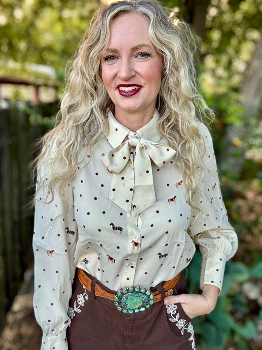 A woman with long blonde hair wearing a cream-colored long-sleeve blouse with a bow collar and small brown polka dots and horse motifs, paired with brown pants and a turquoise concho belt, standing outdoors in front of a blurred green background.
