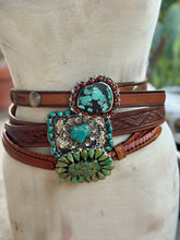 A multi-strand brown leather belts with western-style buckles, displayed on a cream-colored mannequin torso. The belt features a large silver-toned concho buckle with turquoise and black stones, a smaller oval turquoise buckle, and braided leather bands.