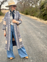 Woman in a patterned coat and blue jeans standing on a road with trees in the background