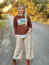 Woman wearing a brown sweatshirt with a horse graphic and floral pants standing on a road.